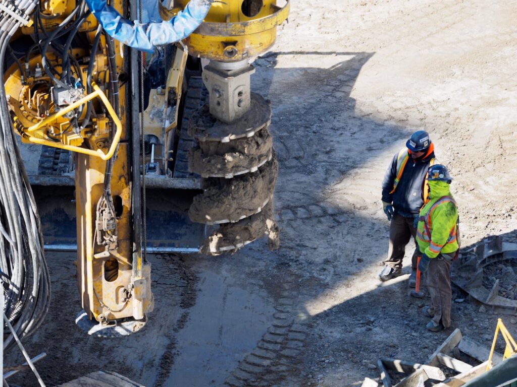 Two workers in protective gear at a construction site with heavy drilling machinery.